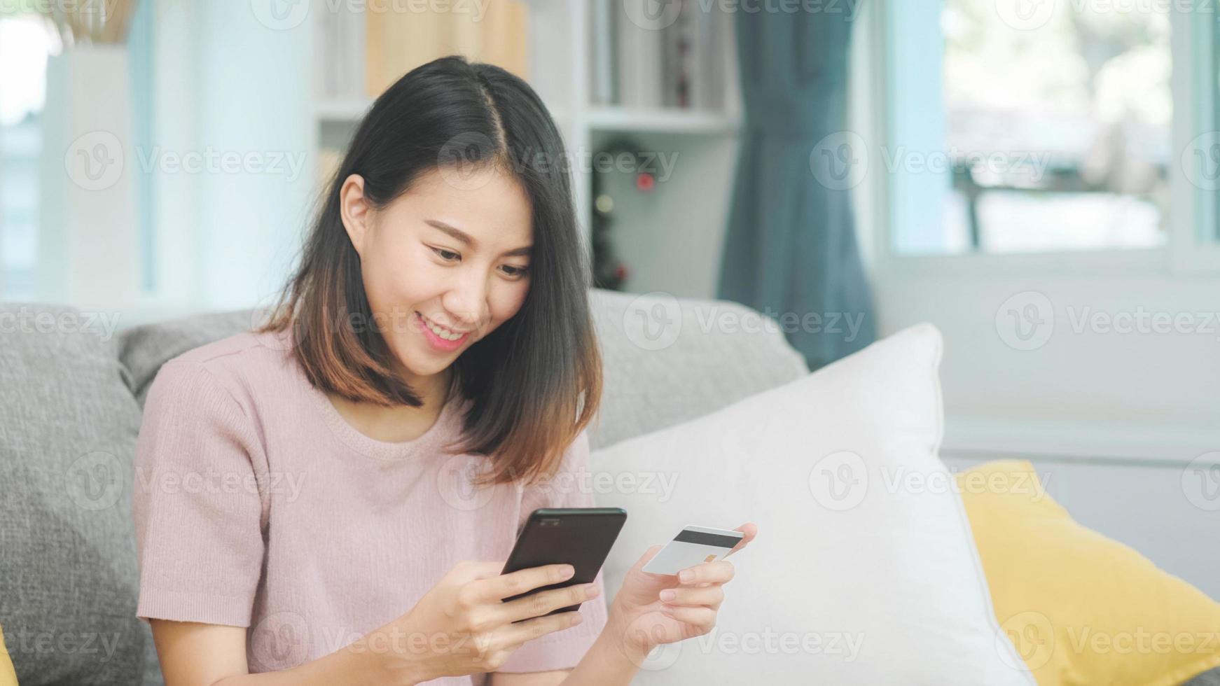 Joven mujer asiática sonriente con smartphone comprando compras en línea con tarjeta de crédito mientras está acostado en el sofá cuando se relaja en la sala de estar en casa. estilo de vida de las mujeres de etnia latina e hispana en el concepto de casa. foto