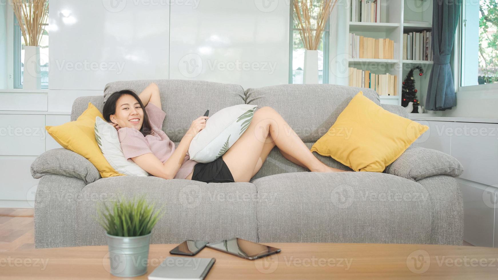 Joven mujer asiática adolescente viendo la televisión en casa, mujer sintiéndose feliz acostado en el sofá en la sala de estar. mujer de estilo de vida relajarse por la mañana en el concepto de hogar. foto