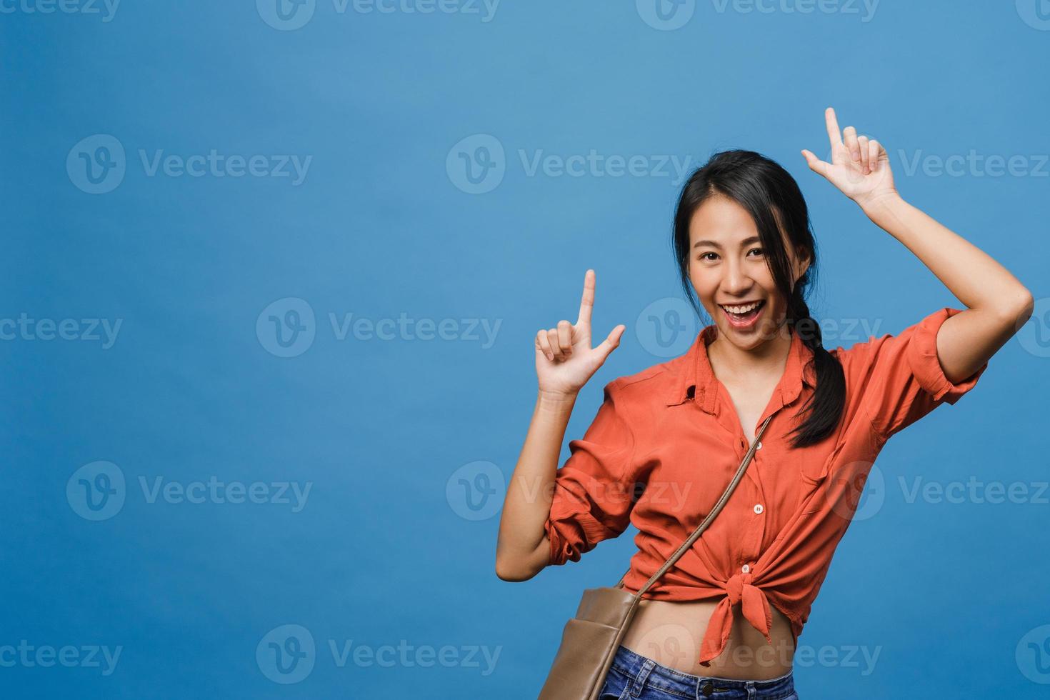 Portrait of young Asian lady smiling with cheerful expression, shows something amazing at blank space in casual cloth and looking at camera isolated over blue background. Facial expression concept. photo