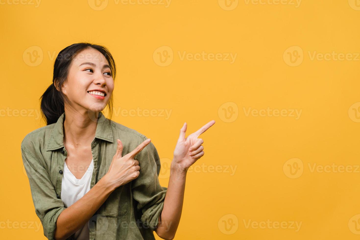 Portrait of young Asian lady smiling with cheerful expression, shows something amazing at blank space in casual clothing and standing isolated over yellow background. Facial expression concept. photo