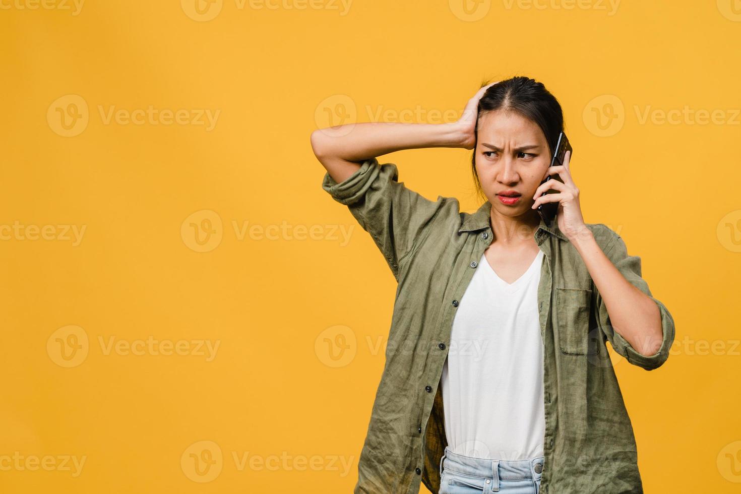 jovencita asiática habla por teléfono con expresión negativa, gritos emocionados, gritos emocionales enojados en ropa casual y se para aislado sobre fondo amarillo con espacio de copia en blanco. concepto de expresión facial. foto