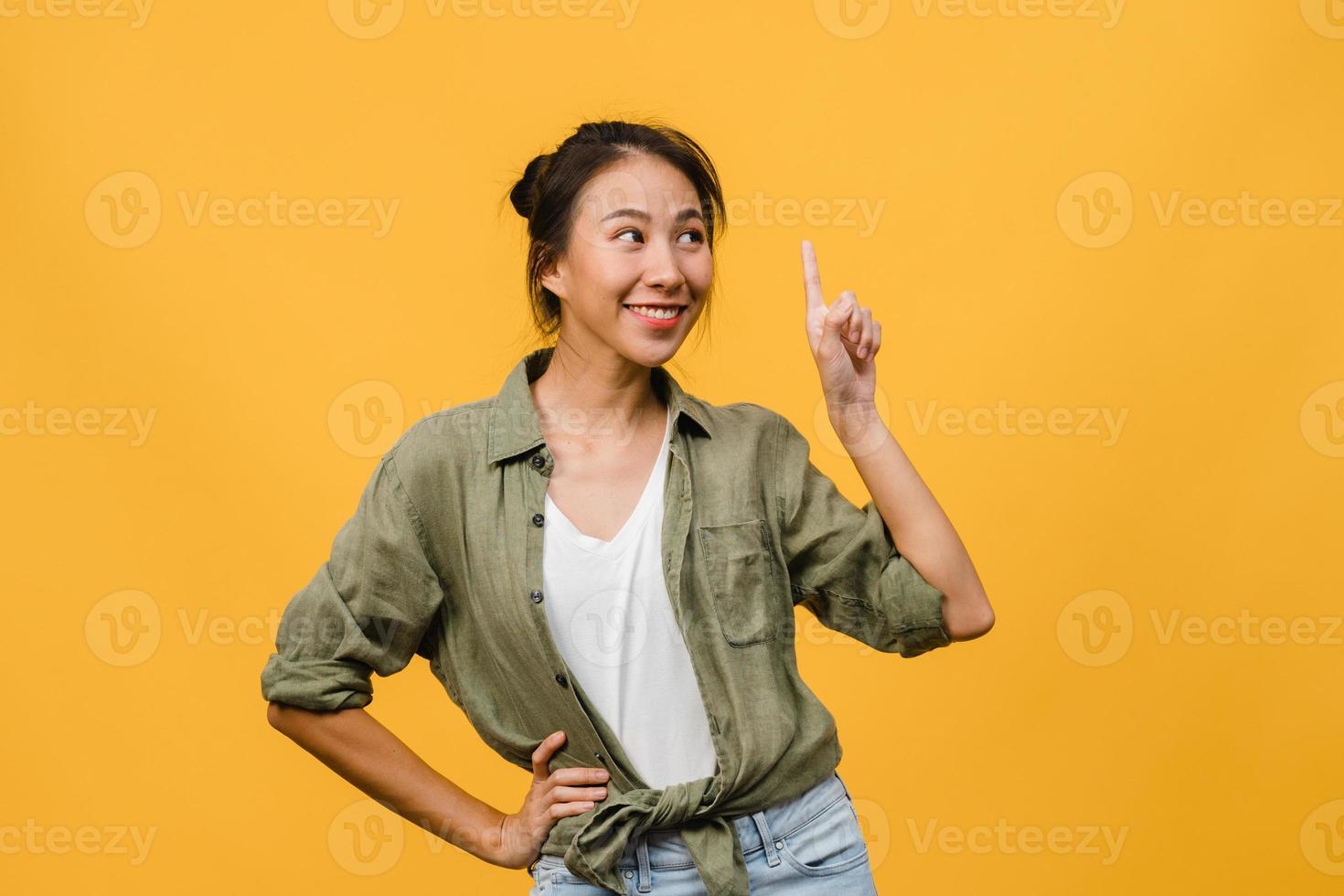 Portrait of young Asian lady smiling with cheerful expression, shows something amazing at blank space in casual clothing and standing isolated over yellow background. Facial expression concept. photo