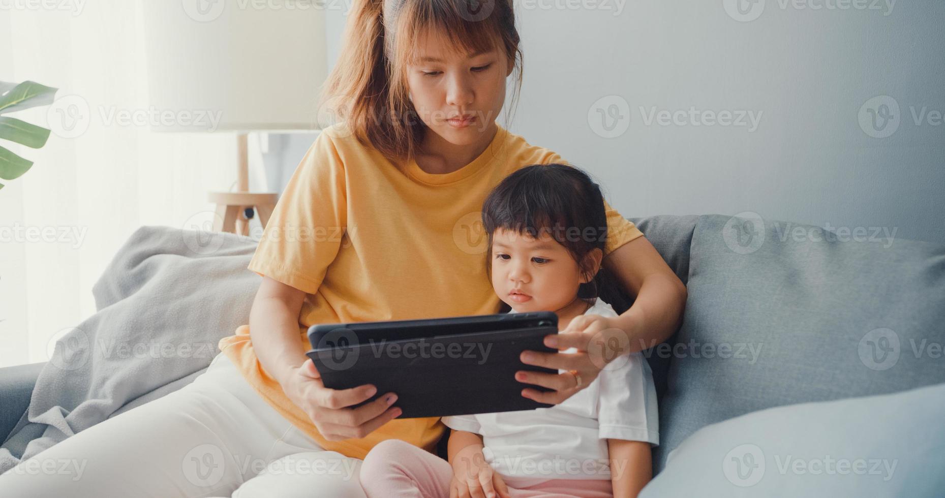 Feliz madre de familia asiática alegre y niño lindo usando dibujos animados de interés de tableta digital y ver películas divirtiéndose relajarse en el sofá en la sala de estar de la casa. pasar tiempo juntos, cuarentena por coronavirus. foto