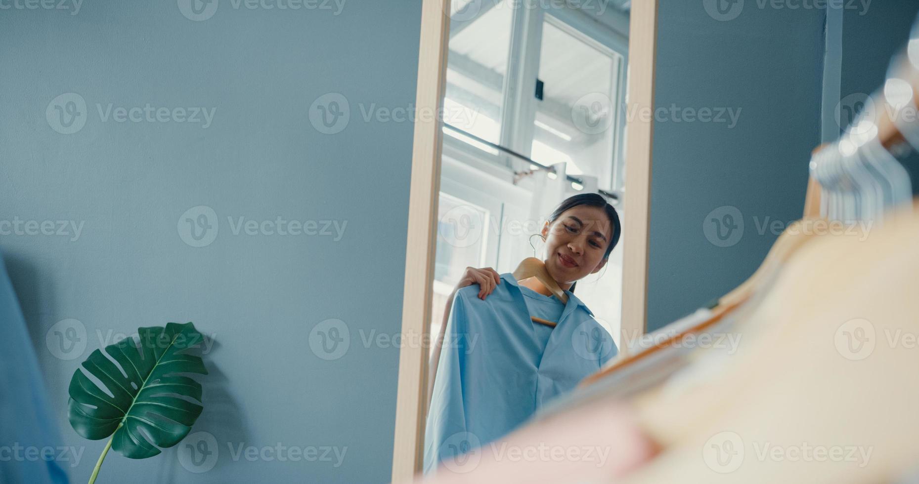 hermosa dama atractiva de asia elegir ropa en perchero vestirse mirando en el espejo en la sala de estar en casa. chica piensa qué ponerse camisa casual. las mujeres de estilo de vida se relajan en el concepto de hogar. foto