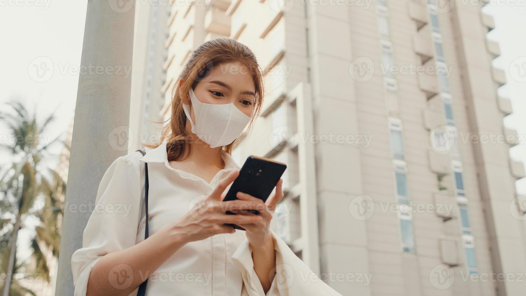 Exitosa empresaria asiática joven en ropa de oficina de moda con mascarilla médica con teléfono inteligente mientras camina solo al aire libre en la ciudad moderna urbana por la mañana. concepto de negocio en movimiento. foto