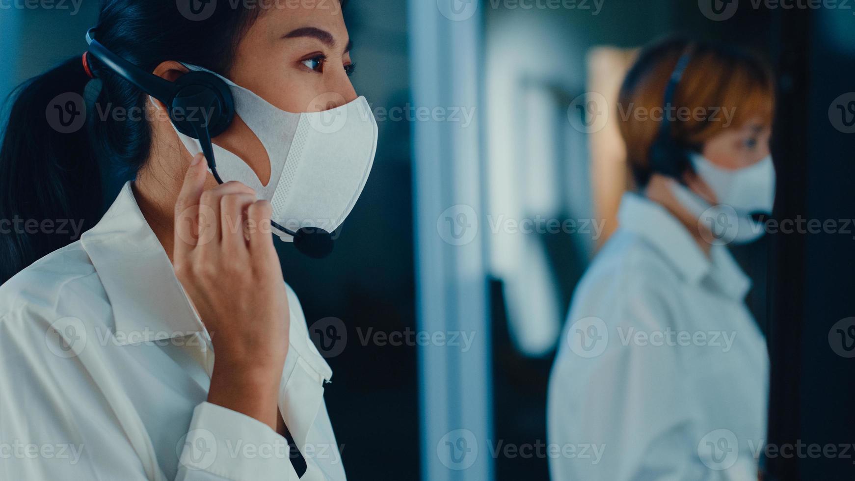 Millennial Asia young call center team or customer support service executive wearing face mask prevent covid-19 using computer and microphone headset working technical support in late night office. photo