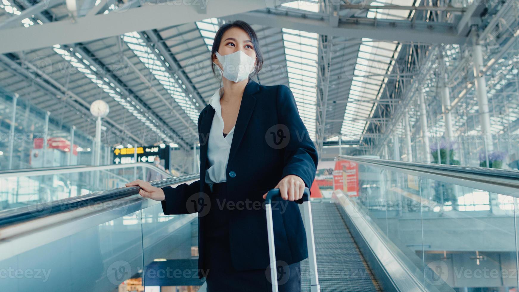 chica de negocios asiática usa mascarilla arrastre soporte de equipaje en la escalera mecánica mire alrededor camine hasta la terminal en el aeropuerto internacional. pandemia de covid viajero de negocios, concepto de distanciamiento social de viajes de negocios. foto