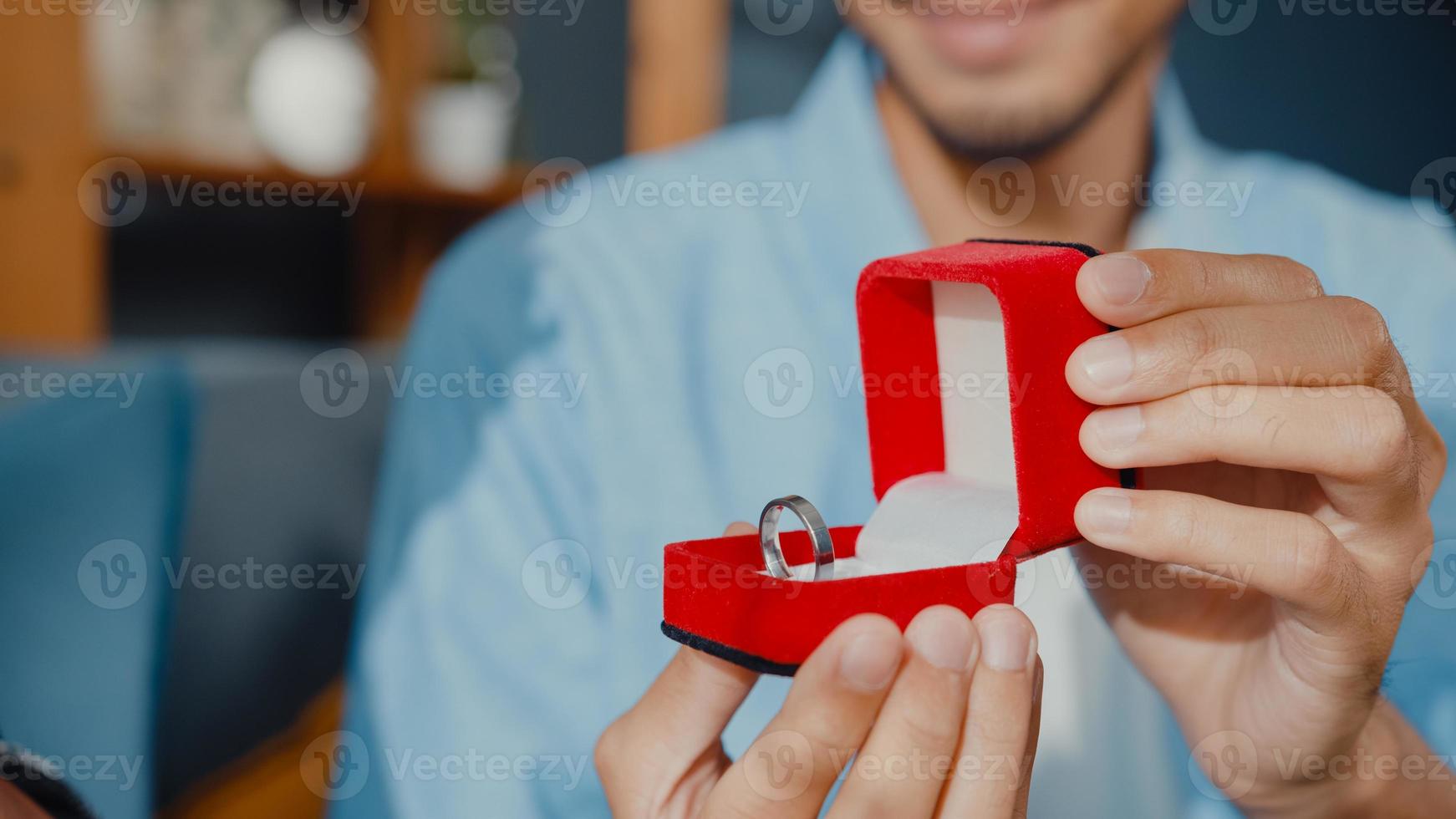 Close up happy asian couple man and woman smiling boyfriend sit on couch surprise girlfriend with engagement ring in red box ask her for a marriage proposal at home. Anniversary romantic concept. photo