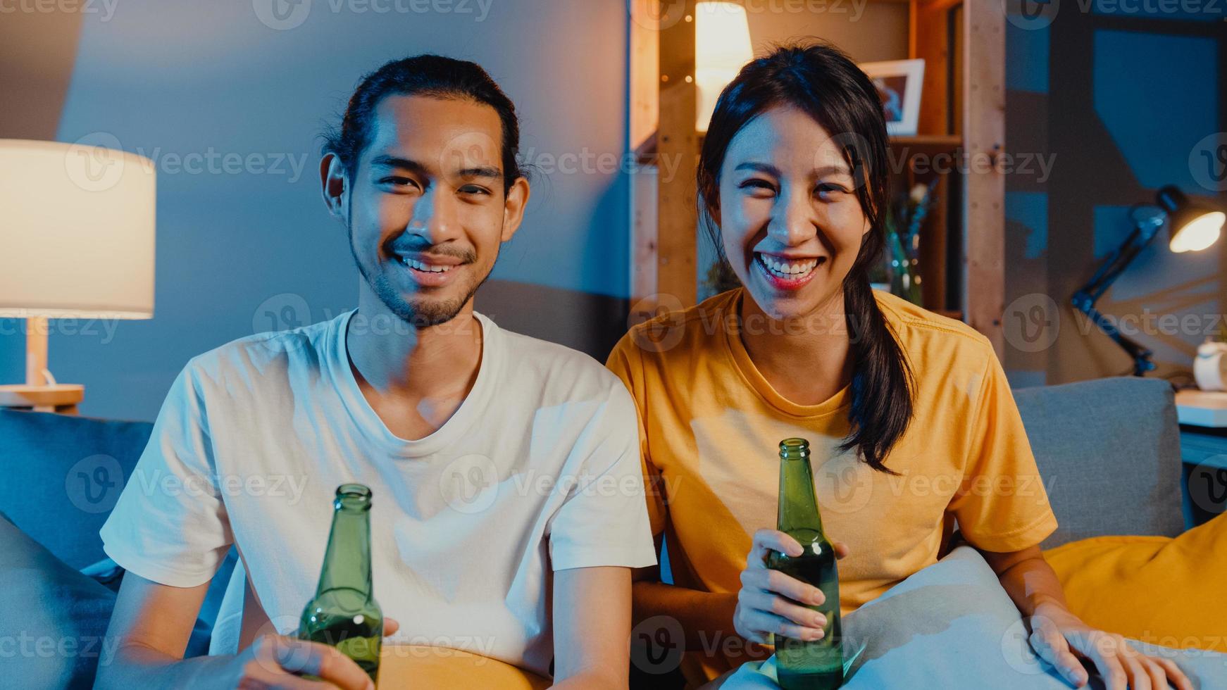 Happy young asian couple looking at camera enjoy night party event online sit couch video call with friends toast drink beer via video call online in living room at home, Social distancing concept. photo
