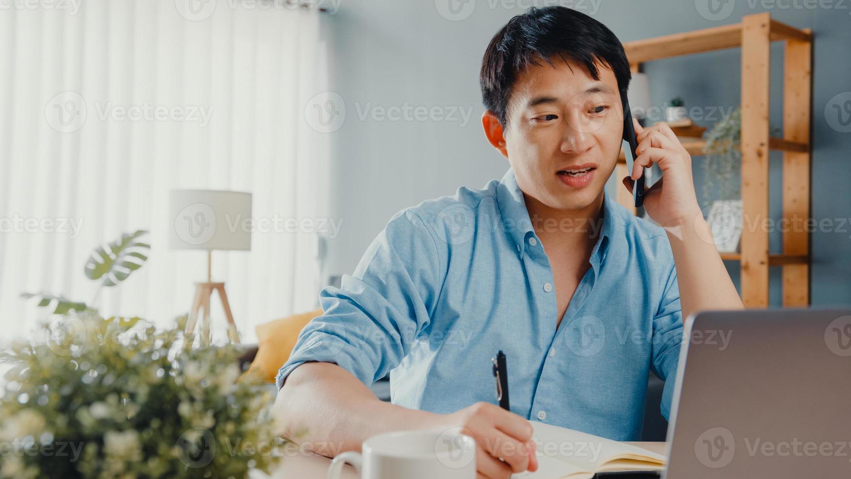 Freelance Asia guy casual wear using laptop talk on cell phone in living room at house. Working from home, remotely work, distance education, social distancing, quarantine for corona virus prevention. photo