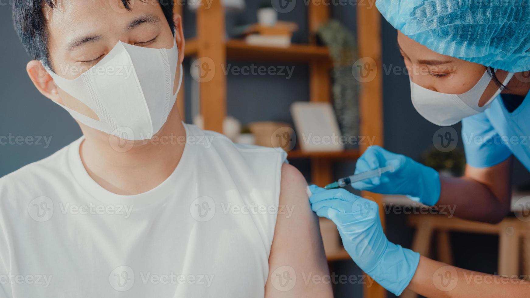 Young Asia lady nurse giving Covid-19 or flu antivirus vaccine shot to male patient wear face mask protection from virus disease sit on couch in living room at house. Vaccination concept. photo
