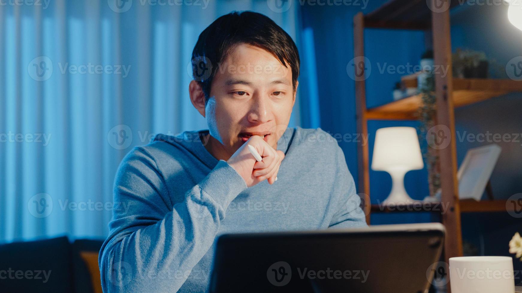 Asia businessman focus online video call meeting assignment on paperwork with colleague in tablet computer in living room at home overtime at night, Work from home coronavirus pandemic concept. photo