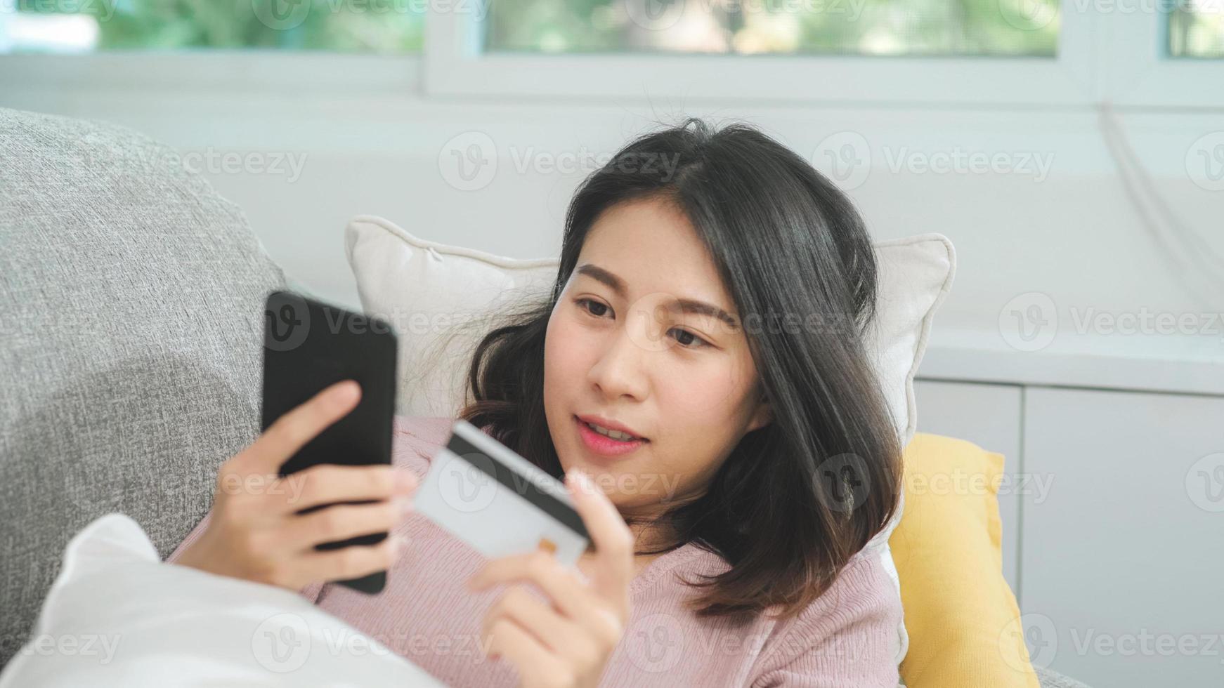 Joven mujer asiática sonriente con smartphone comprando compras en línea con tarjeta de crédito mientras está acostado en el sofá cuando se relaja en la sala de estar en casa. estilo de vida de las mujeres de etnia latina e hispana en el concepto de casa. foto