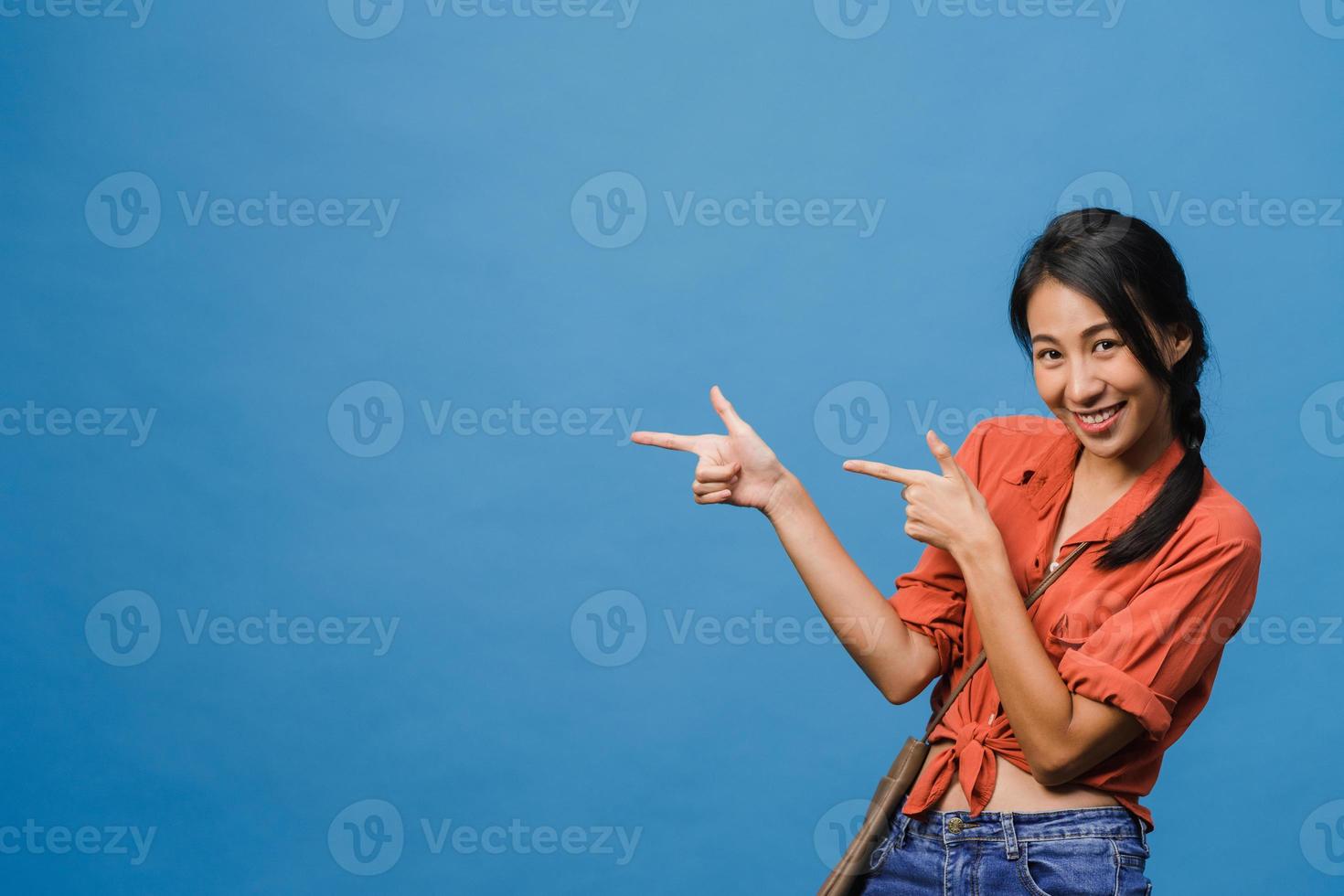 Portrait of young Asian lady smiling with cheerful expression, shows something amazing at blank space in casual cloth and looking at camera isolated over blue background. Facial expression concept. photo