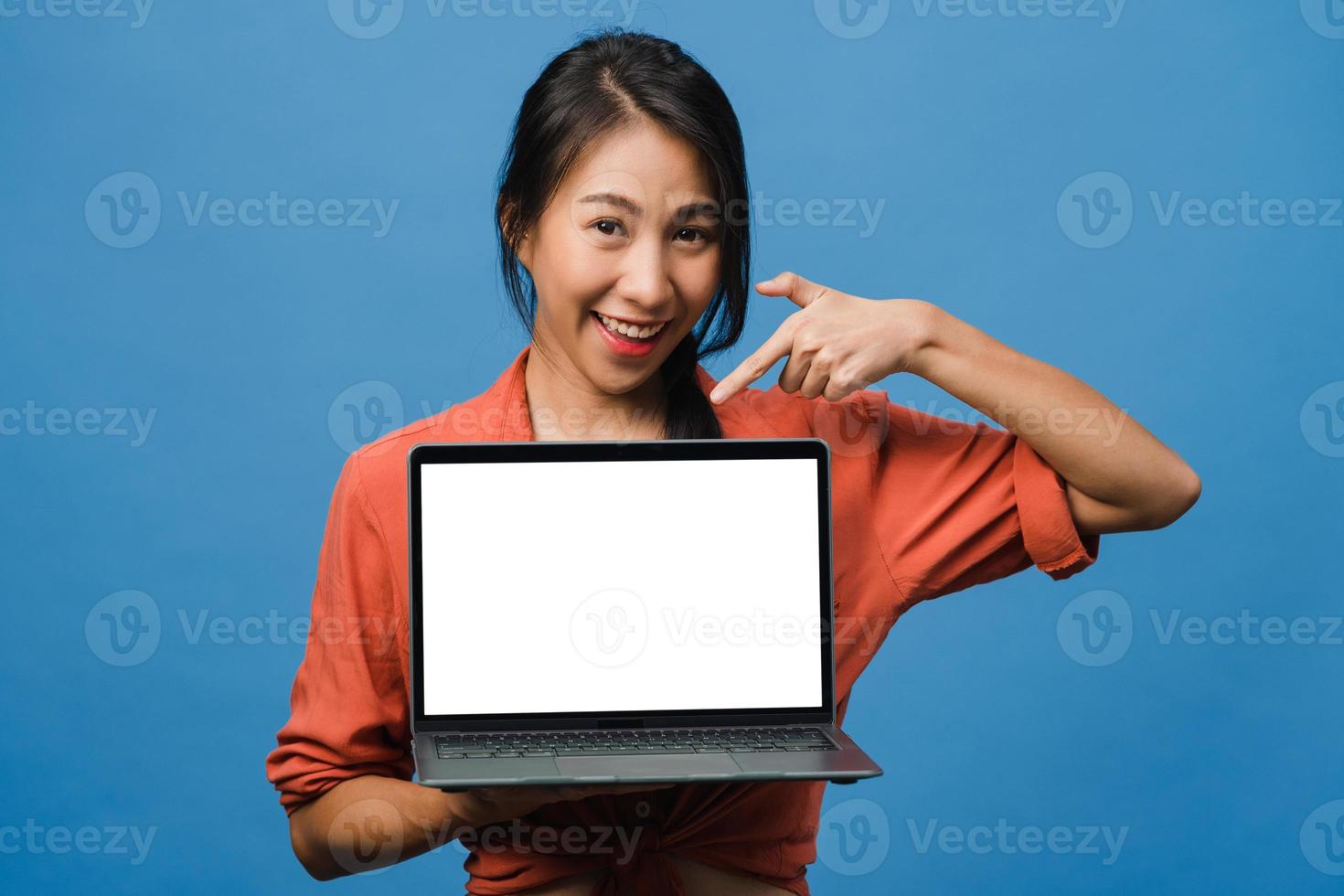 Joven asiática muestra una pantalla de computadora portátil vacía con expresión positiva, sonríe ampliamente, vestida con ropa casual sintiendo felicidad aislada sobre fondo azul. computadora con pantalla blanca en mano femenina. foto