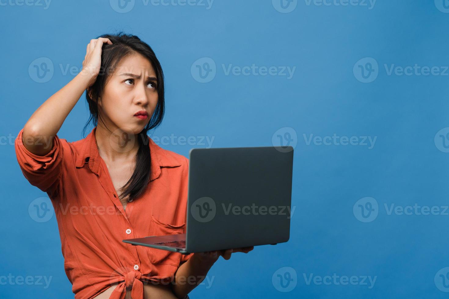 Jovencita asiática usando laptop con expresión negativa, gritando emocionado, llorando emocionalmente enojado en ropa casual y parado aislado sobre fondo azul con espacio de copia en blanco. concepto de expresión facial. foto