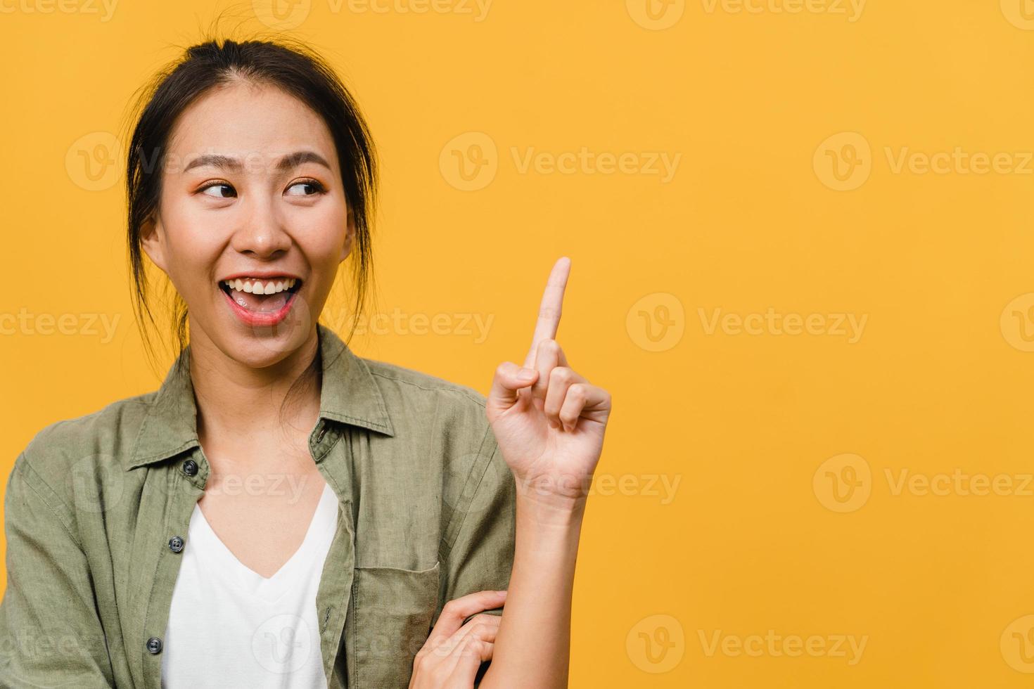 Portrait of young Asian lady smiling with cheerful expression, shows something amazing at blank space in casual clothing and standing isolated over yellow background. Facial expression concept. photo