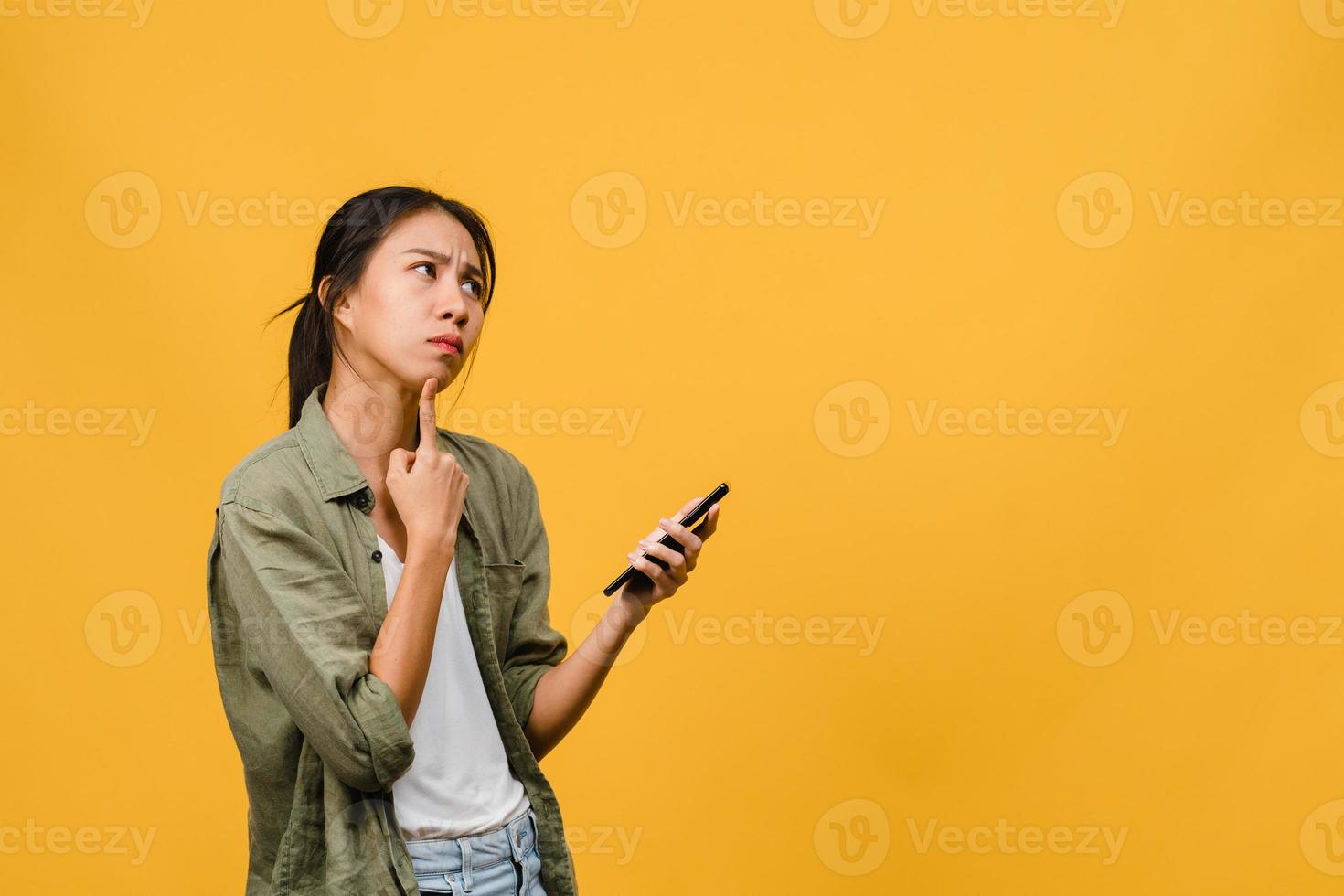 Thinking dreaming young Asia lady using phone with positive expression, dressed in casual cloth feeling happiness and stand isolated on yellow background. Happy adorable glad woman rejoices success. photo