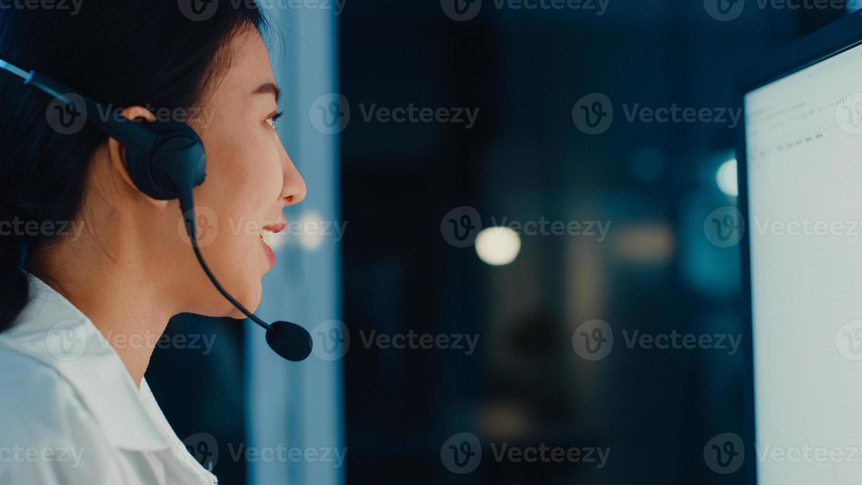 Millennial Asia young call center agent or customer support service executive using computer and microphone headset working technical support in late night office. Telemarketing or sales job concept. photo