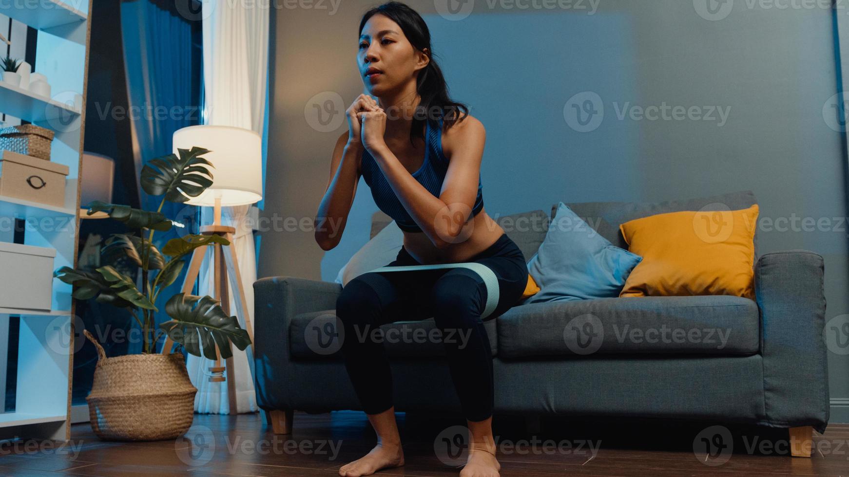 Young Asia lady in sportswear doing squat exercise working out in living room at home at night. Sport and recreation activity, social distancing, quarantine for corona virus prevention concept. photo