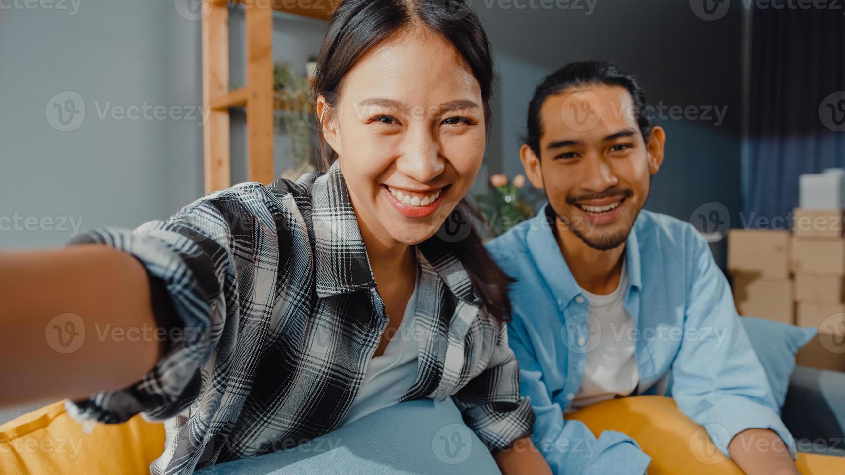 Happy young asian couple man and woman sit couch use looking at camera video call with friends and family in living room at home. Stay at home quarantine, Social distancing, Young married concept. photo