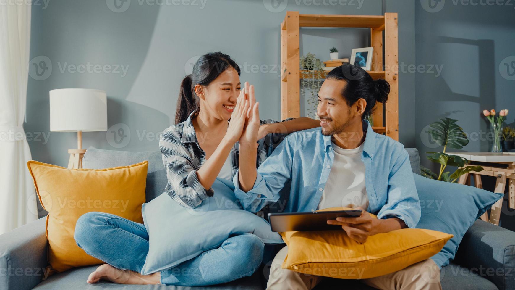 Feliz pareja joven y atractiva asiática, hombre y mujer, sentarse en el sofá, usar la tableta, comprar muebles en línea, decorar el hogar en la sala de estar de la nueva casa. joven casado mudanza concepto online comprador de casa. foto