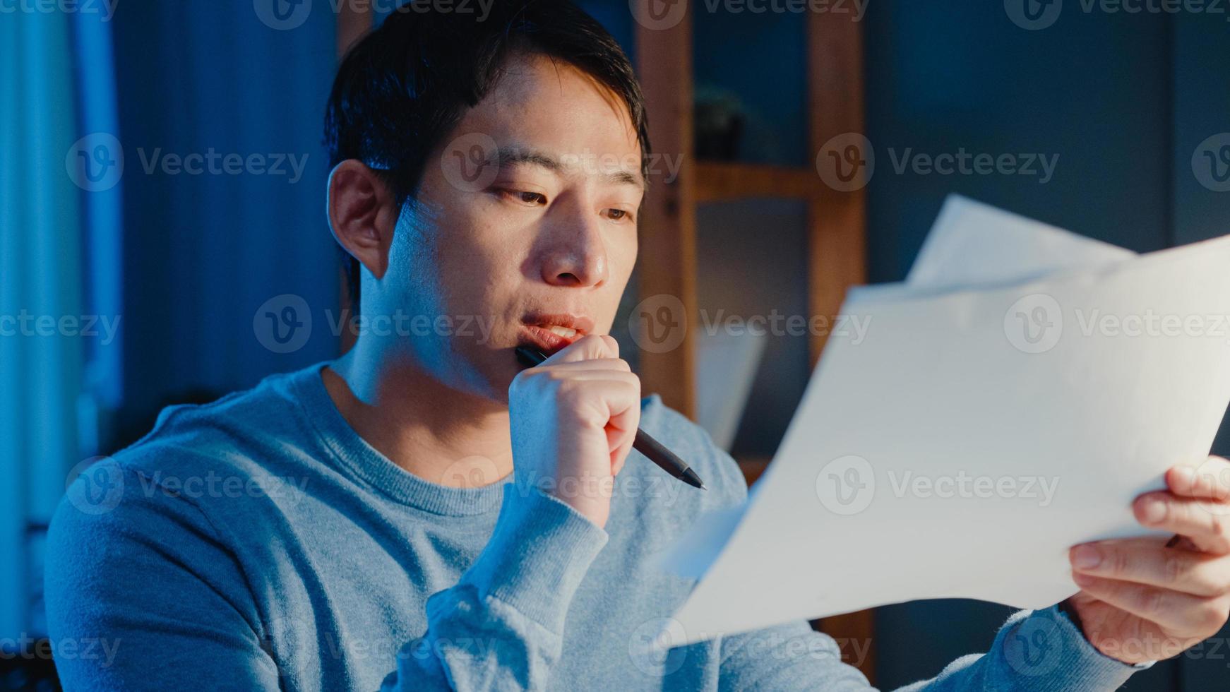 Hombre de negocios autónomo asiático se enfoca en el tipo de trabajo en la computadora portátil ocupado con un gráfico lleno de papeleo en el escritorio en la sala de estar en casa horas extras por la noche, trabaja desde casa durante el concepto de pandemia covid-19. foto