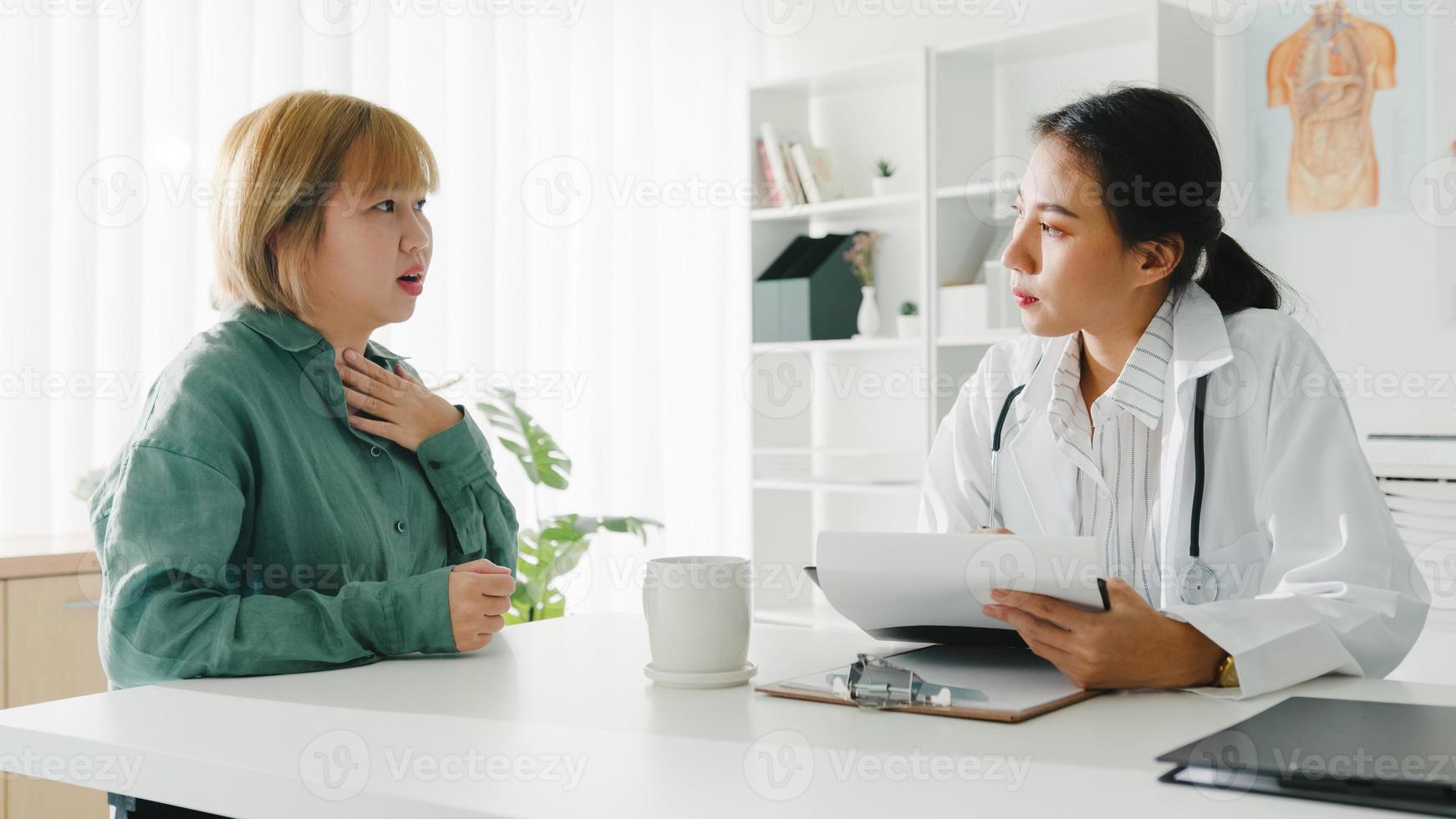 La joven doctora asiática en uniforme médico blanco que usa el portapapeles está brindando una gran charla de noticias sobre los resultados o los síntomas con la paciente sentada en el escritorio en la clínica de salud o en la oficina del hospital. foto