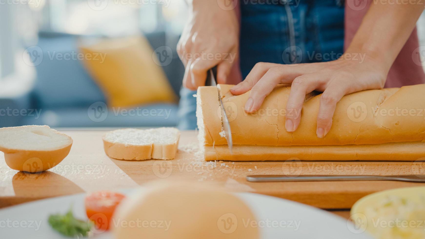 Manos de joven mujer asiática chef sosteniendo un cuchillo cortando pan integral sobre tabla de madera en la mesa de la cocina en casa. producción de pan casero fresco, alimentación saludable y concepto de panadería tradicional. foto