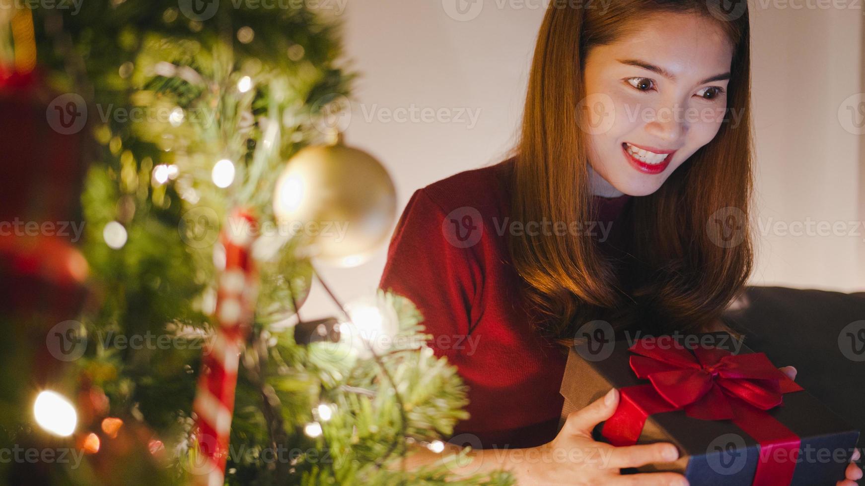 Mujer joven de Asia con videollamada de tableta hablando con pareja con caja de regalo de Navidad, árbol de Navidad decorado con adornos en la sala de estar en casa. noche de navidad y fiesta de año nuevo. foto