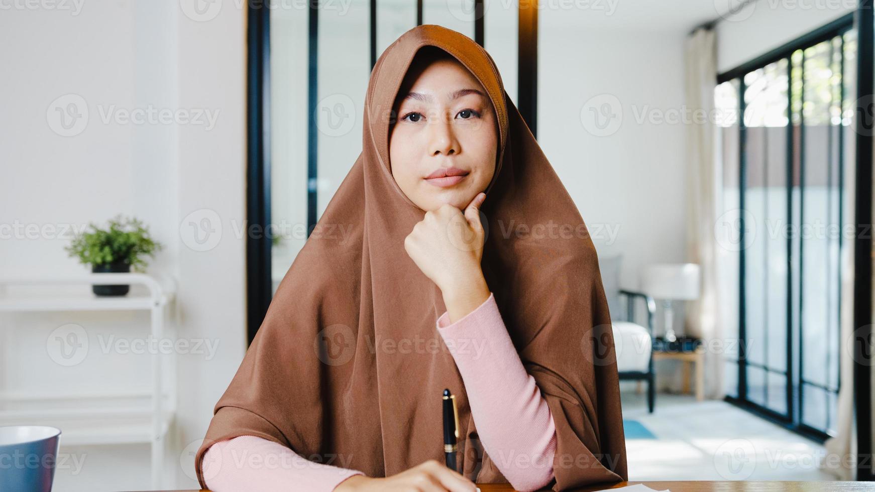 Asia muslim lady wear hijab using computer laptop talk to colleagues about plan in video call meeting while remotely work from home at living room. Social distancing, quarantine for corona virus. photo