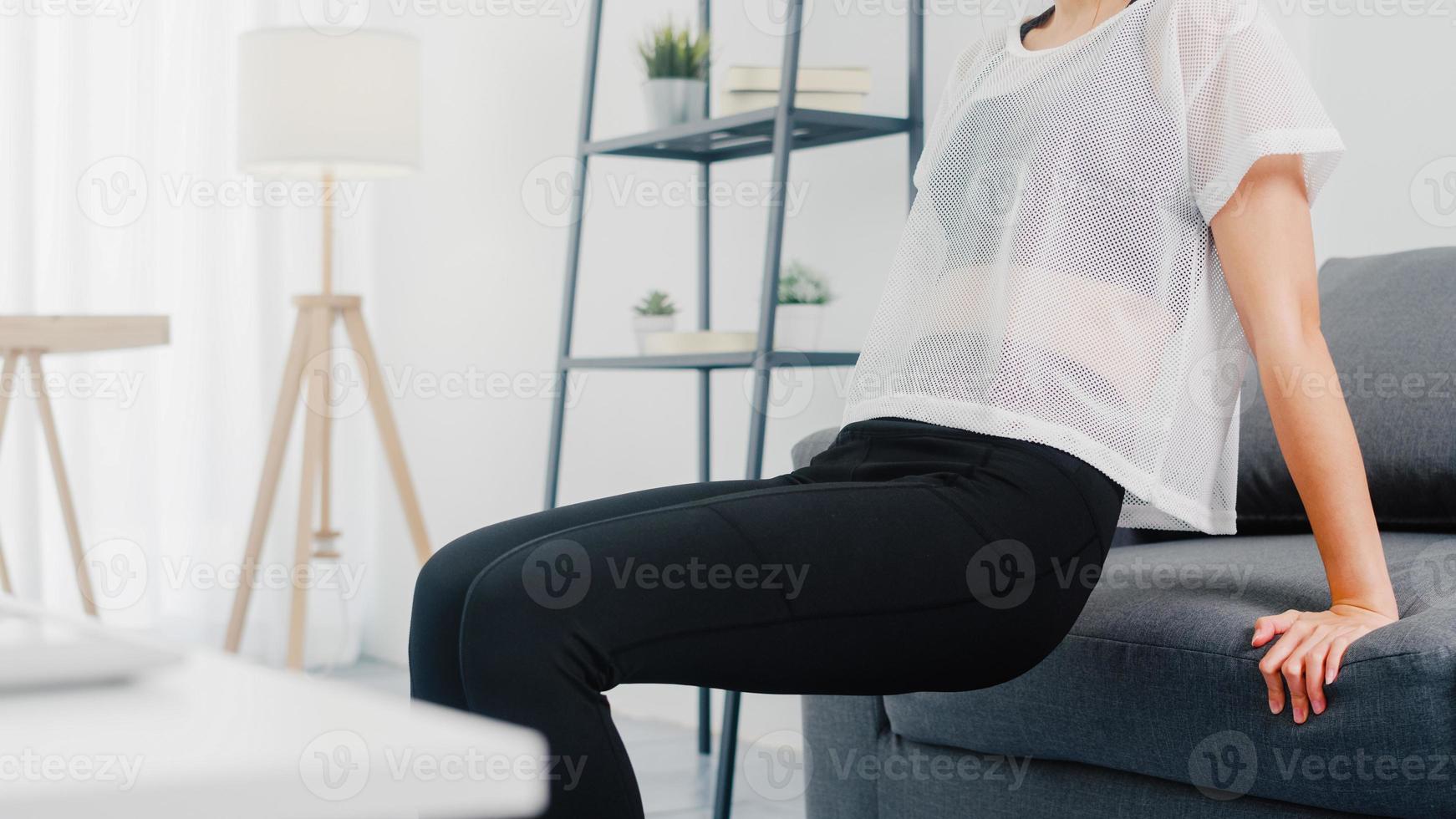 Young Korean lady in sportswear exercises doing working out doing tricep dips leaning on couch in living room at home. Social distance, Isolation during the virus. Exercises for the lower body. photo