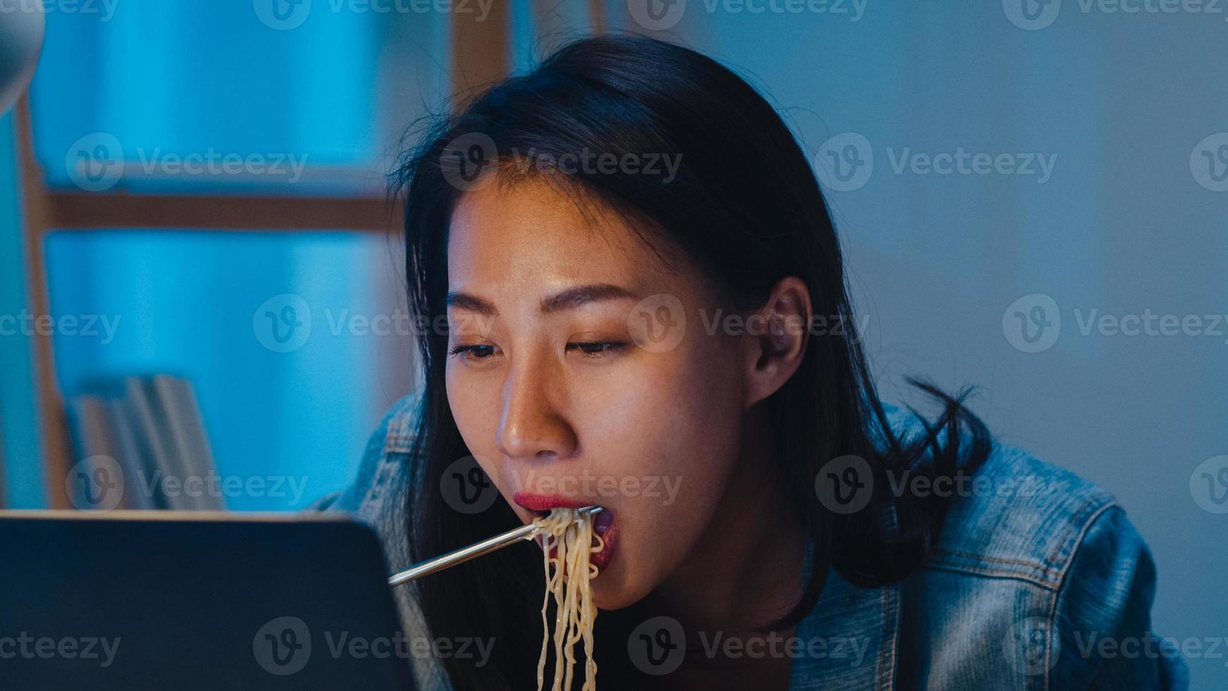 mujeres de negocios inteligentes independientes que comen fideos instantáneos estrés, cansadas mientras trabajan en la computadora portátil en la sala de estar en casa por la noche. feliz joven asiática sentada en el escritorio trabajar horas extras, disfrutar del tiempo de relajación. foto