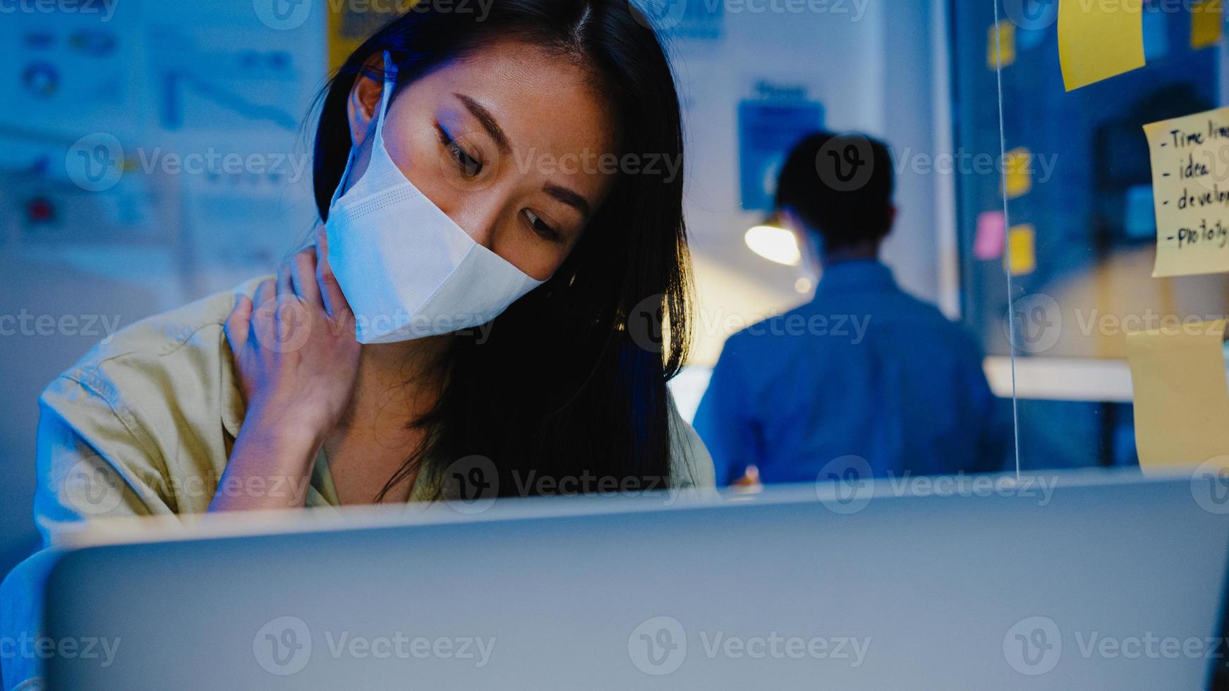 destacó cansado joven asia mujer usar mascarilla usando laptop trabajo duro con síndrome de oficina, dolor de cuello, mientras trabaja horas extras en la oficina. trabajo desde casa sobrecarga nocturna, distanciamiento social. foto