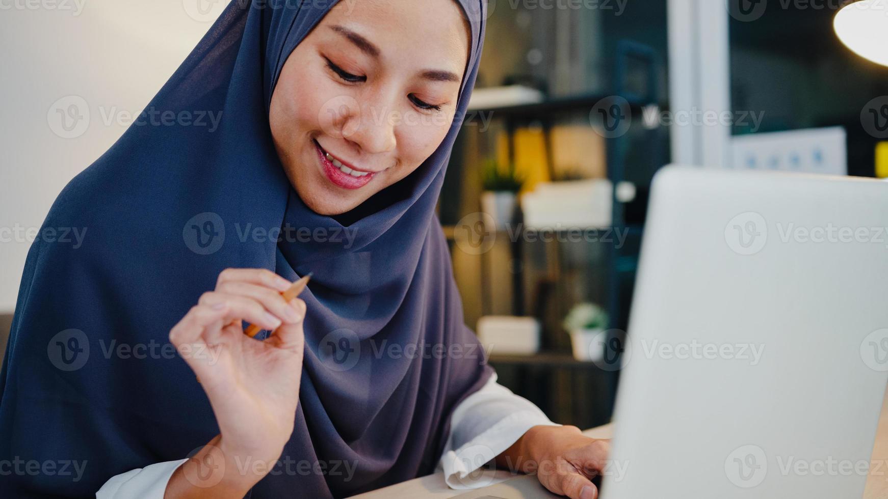 Beautiful Asia muslim lady in headscarf casual wear using laptop in living room at night house. Remotely work from home, new normal lifestyle, social distance, quarantine for corona virus prevention. photo