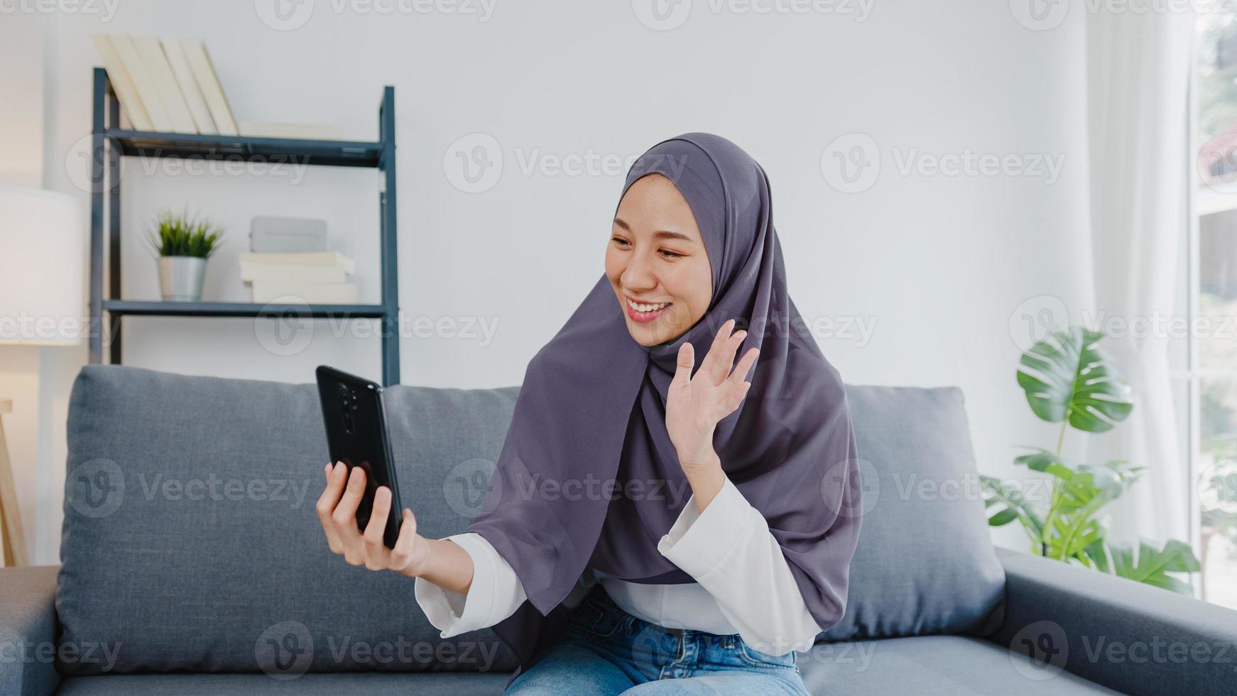 Asia dama musulmana usa hijab usando videollamada telefónica hablando con pareja en casa. joven adolescente haciendo video de vlog en las redes sociales en el sofá de la sala de estar. distanciamiento social, cuarentena por coronavirus. foto