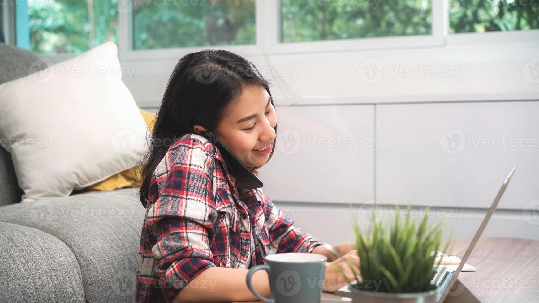 Freelance Asian woman working at home, business female working on laptop and using mobile phone talking with customer on sofa in living room at home. Lifestyle women working at home concept. photo