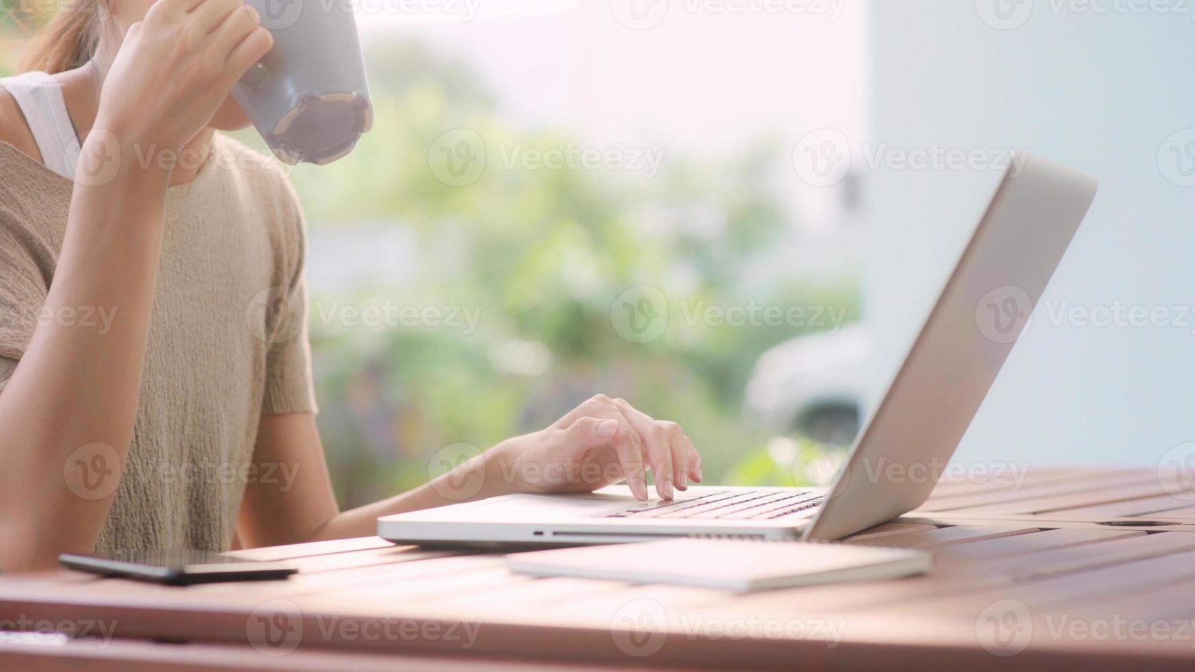 Freelance Asian woman working at home, business female working on laptop and drinking coffee sitting on table in the garden in morning. Lifestyle women working at home concept. photo