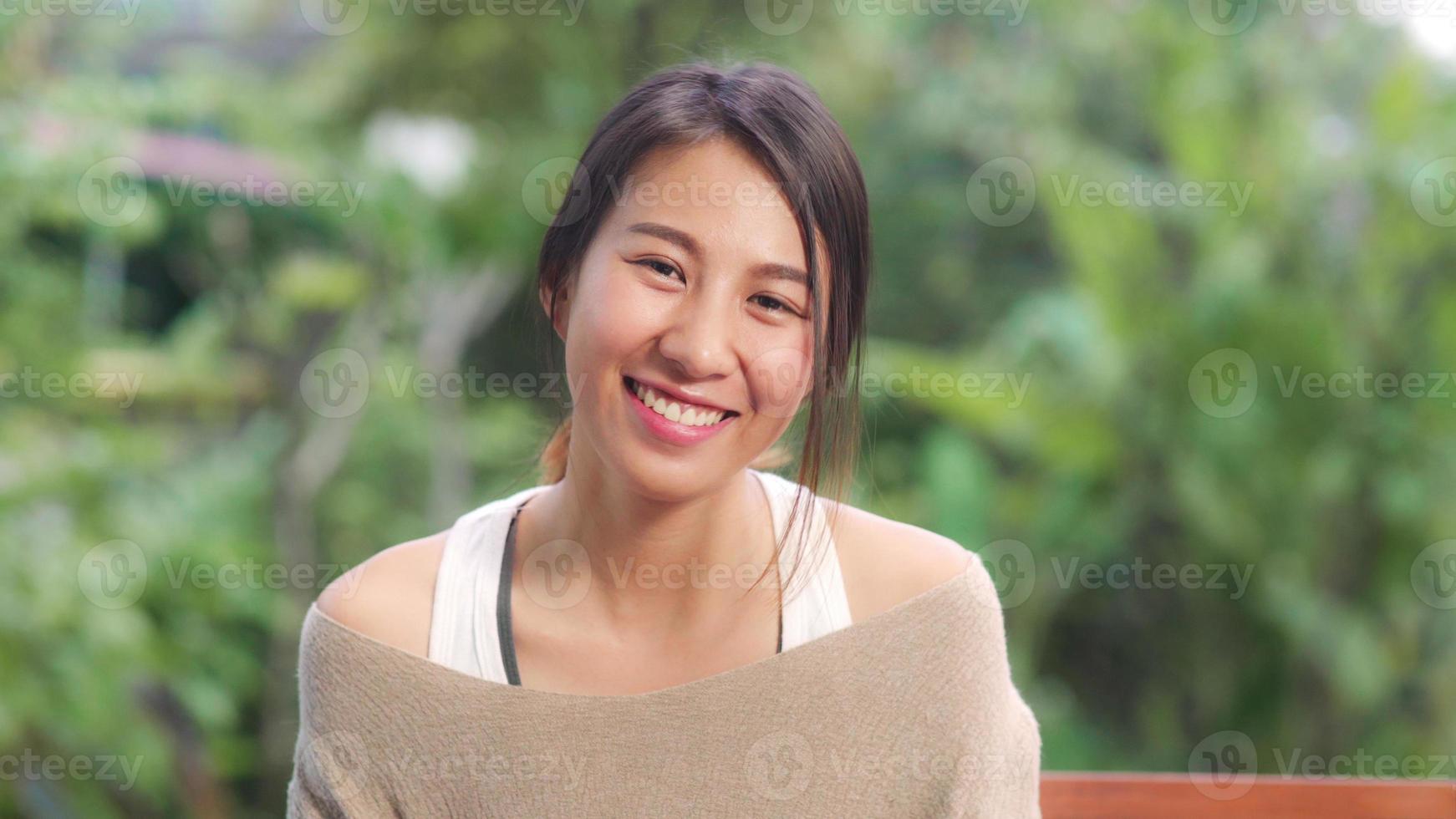 Mujer asiática que se siente feliz sonriendo y mirando a la cámara mientras se relaja en la mesa en el jardín en casa por la mañana. las mujeres de estilo de vida se relajan en el concepto de hogar. foto