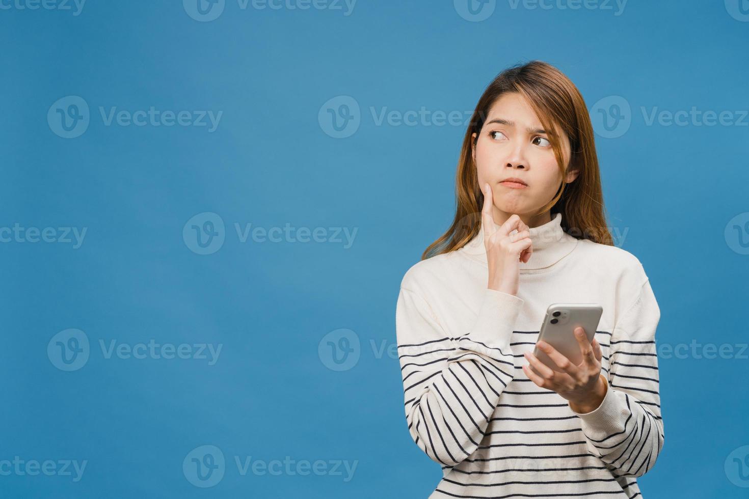 Pensando soñando joven asiática usando el teléfono con expresión positiva, vestida con ropa casual sintiendo felicidad y parada aislada sobre fondo azul. feliz adorable mujer alegre se regocija con el éxito. foto