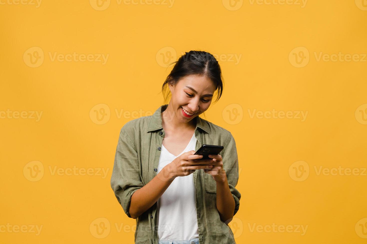 Jovencita asiática que usa el teléfono con expresión positiva, sonríe ampliamente, vestida con ropa casual sintiendo felicidad y parada aislada sobre fondo amarillo. feliz adorable mujer alegre se regocija con el éxito. foto