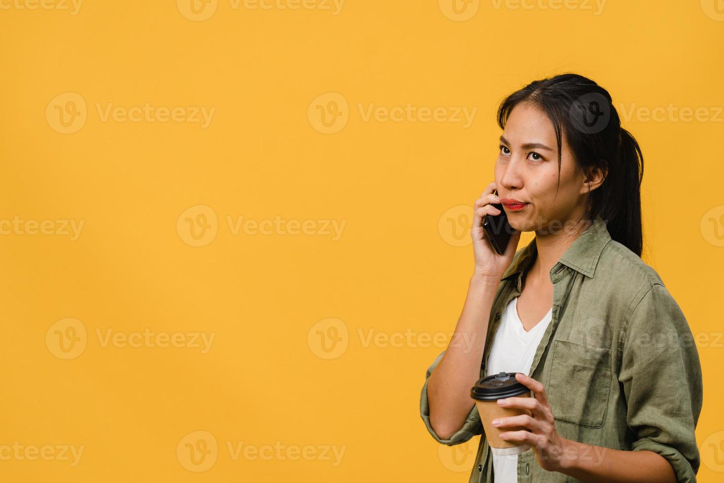 Jovencita asiática habla por teléfono y sostiene la taza de café con expresión negativa, gritos emocionados, llora enojado emocionalmente en ropa casual y se para aislado sobre fondo amarillo. concepto de expresión facial. foto