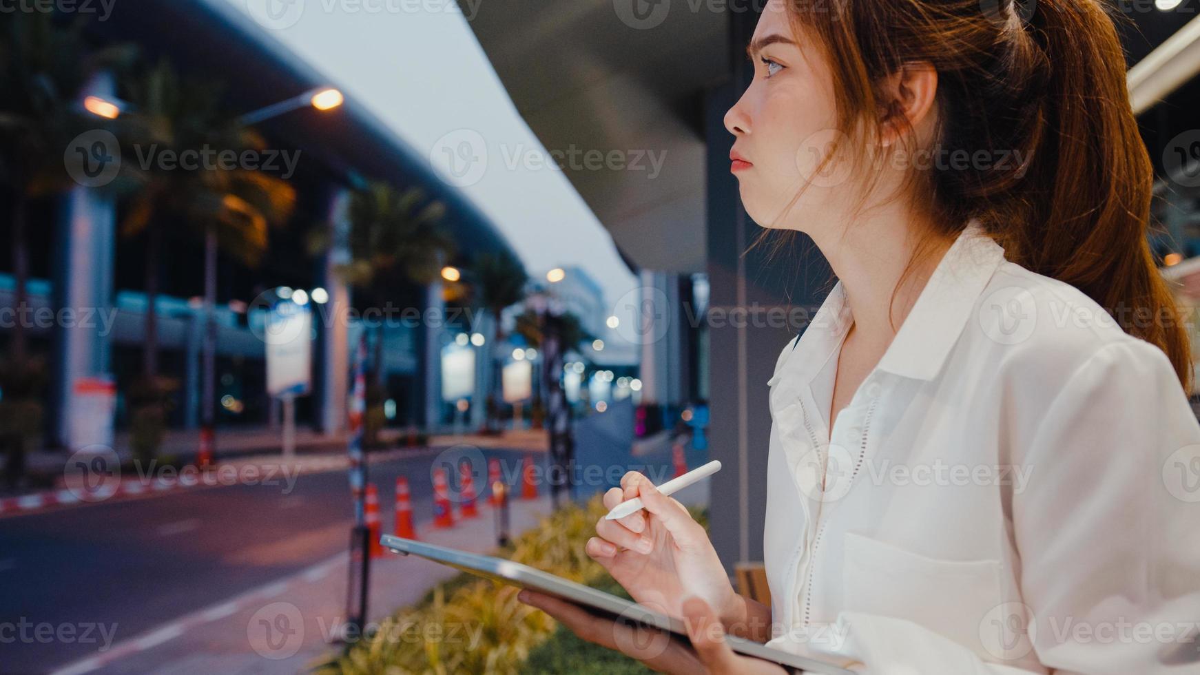 Exitosa empresaria asiática joven en ropa de oficina de moda utilizando tecnología de lápiz inteligente para escribir en tableta digital mientras se sienta sola al aire libre en la ciudad moderna urbana por la noche. concepto de negocio en movimiento. foto