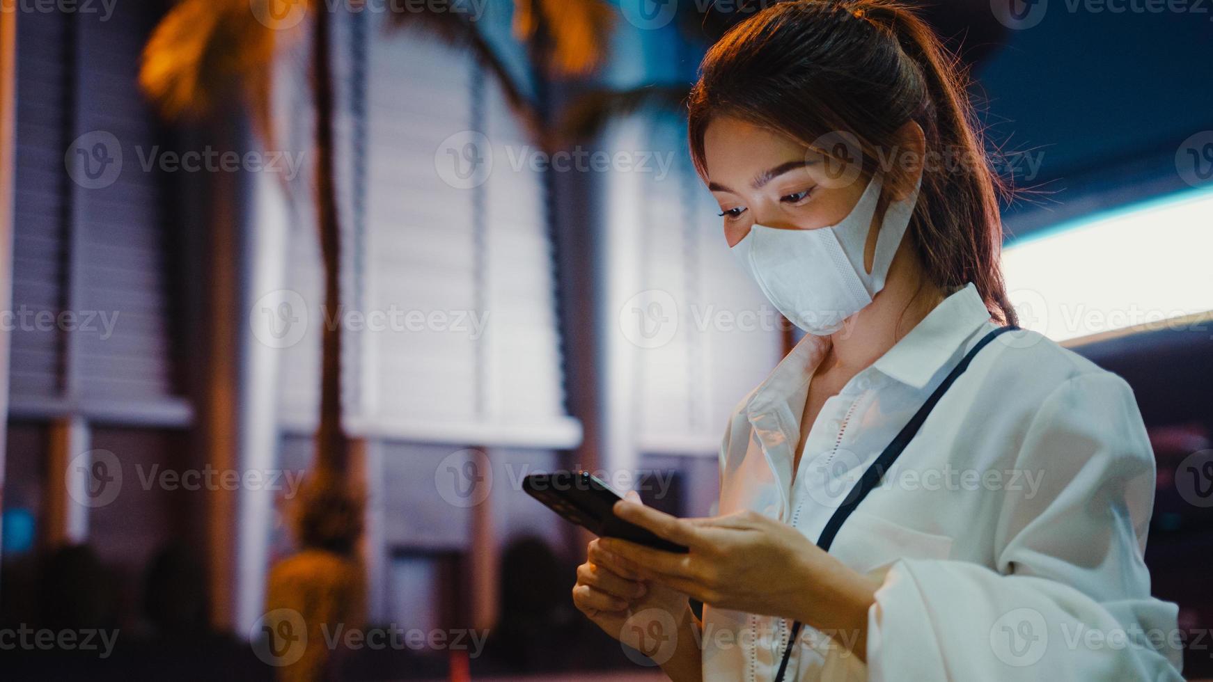 Young Asia businesswoman in fashion office clothes wearing medical face mask using smart phone typing text message while stand outdoors in urban modern city at night. Business on the go concept. photo