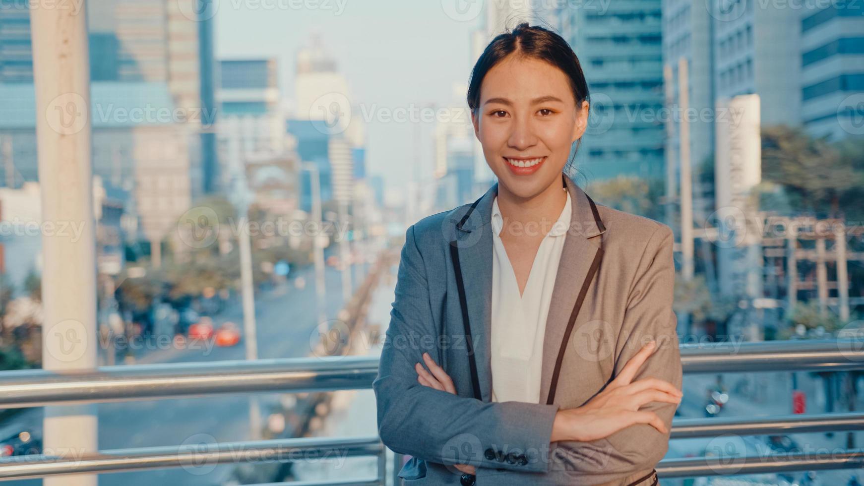 Successful young Asia businesswoman in fashion office clothes smiling and looking at camera while happy standing alone outdoors in urban modern city in the morning. Business on the go concept. photo