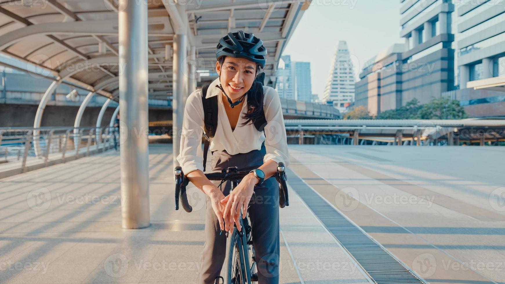 Asian businesswoman go to work at office stand and smiling wear backpack look at camera with bicycle on street around building on a city. Bike commuting, Commute on bike, Business commuter concept. photo