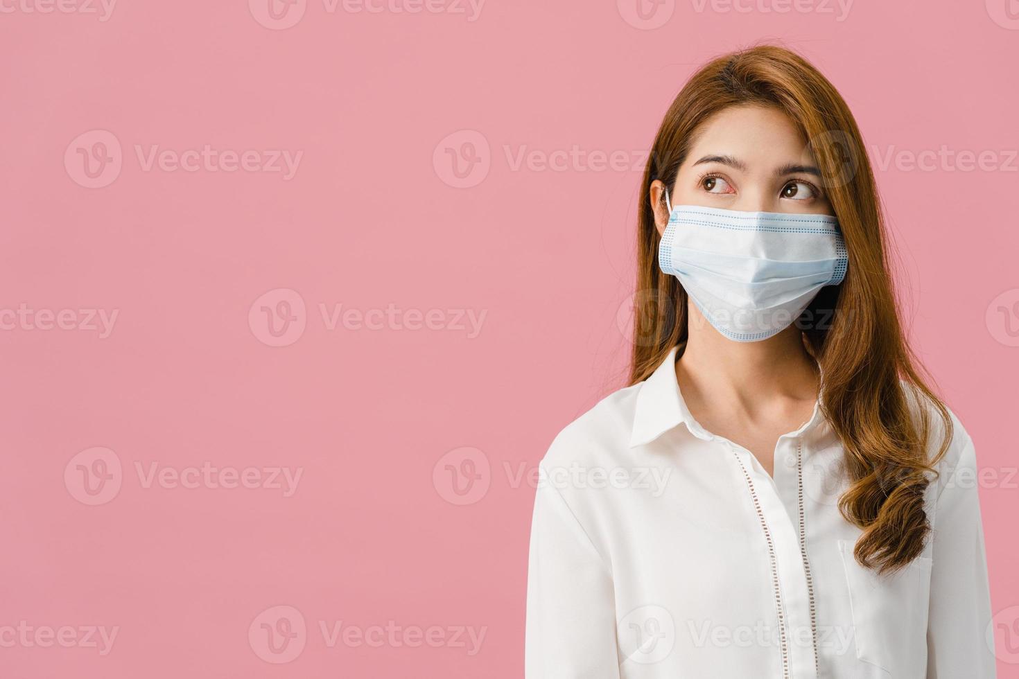 joven asiática con mascarilla médica vestida con un paño informal y mirando el espacio en blanco aislado sobre fondo rosa. autoaislamiento, distanciamiento social, cuarentena para la prevención del virus corona foto