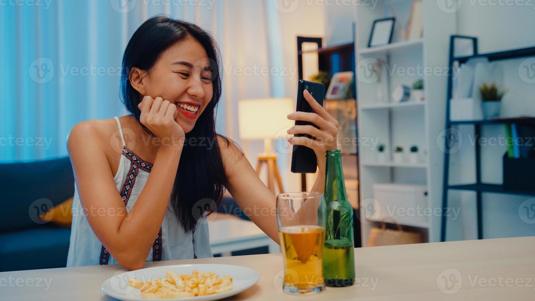 Young Asia lady drinking beer having fun happy moment night party New Year event online celebration via video call by phone at home at night. Social distancing, quarantine for coronavirus prevention. photo