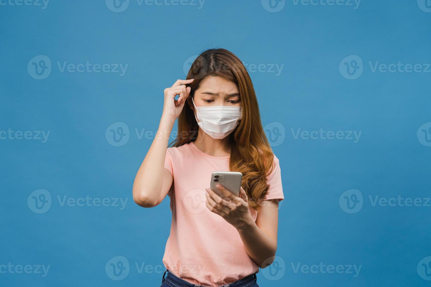 Young Asia girl wearing medical face mask using mobile phone with dressed in casual clothing isolated on blue background. Self-isolation, social distancing, quarantine for corona virus prevention. photo