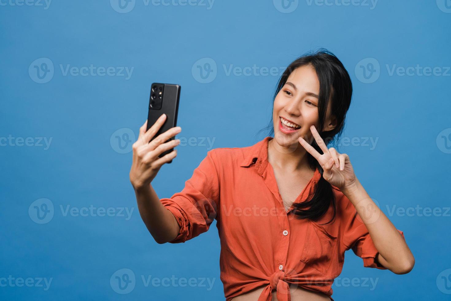 Smiling adorable Asian female making selfie photo on smart phone with positive expression in casual clothing and stand isolated on blue background. Happy adorable glad woman rejoices success.